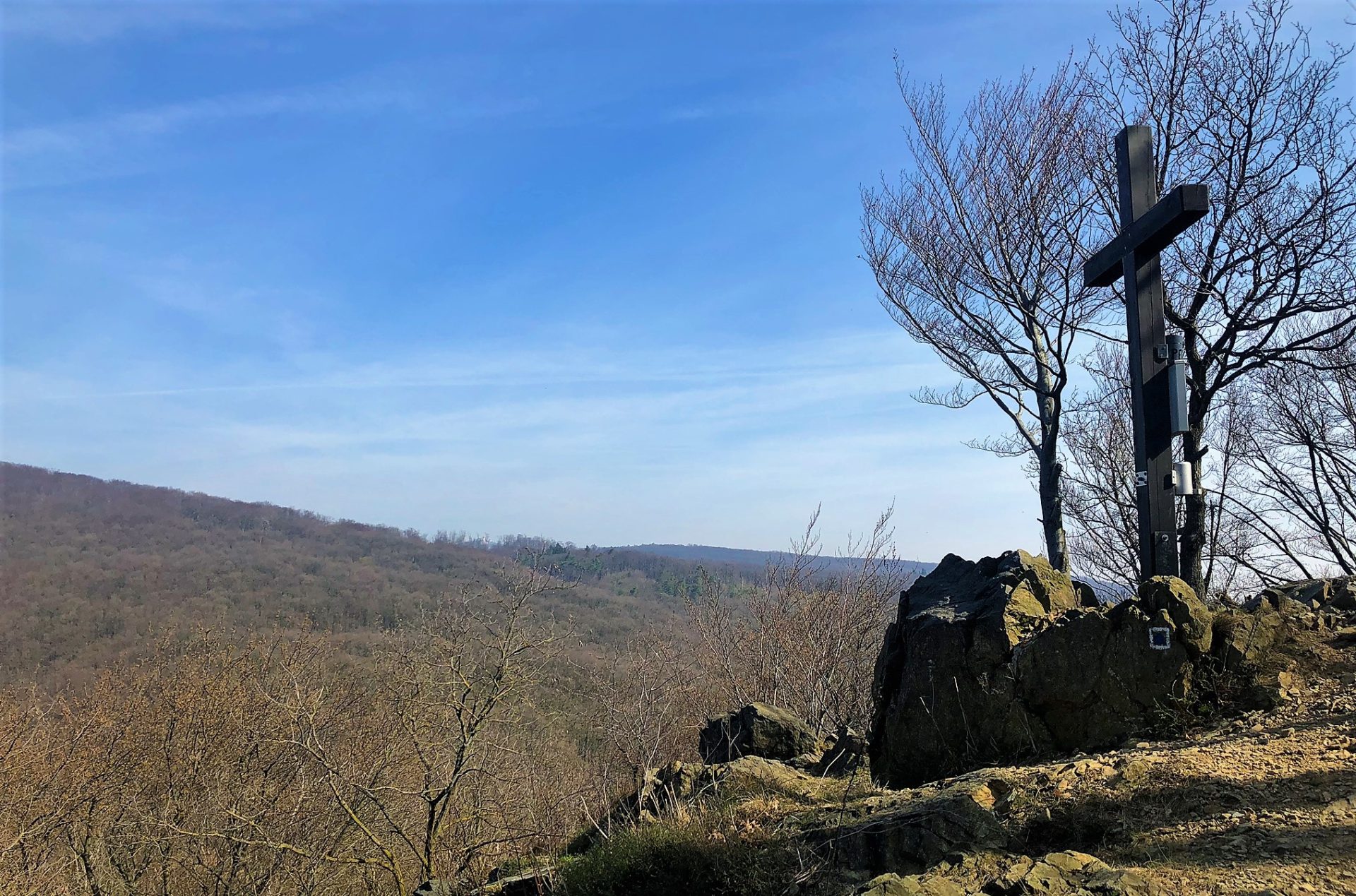 A wooden cross stands on a rocky overlook above a forested valley