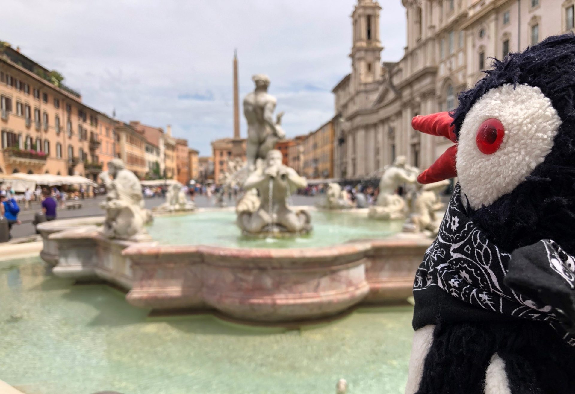 Plush penguin Miško wearing a bandana in front of a fountain in Piazza Navona