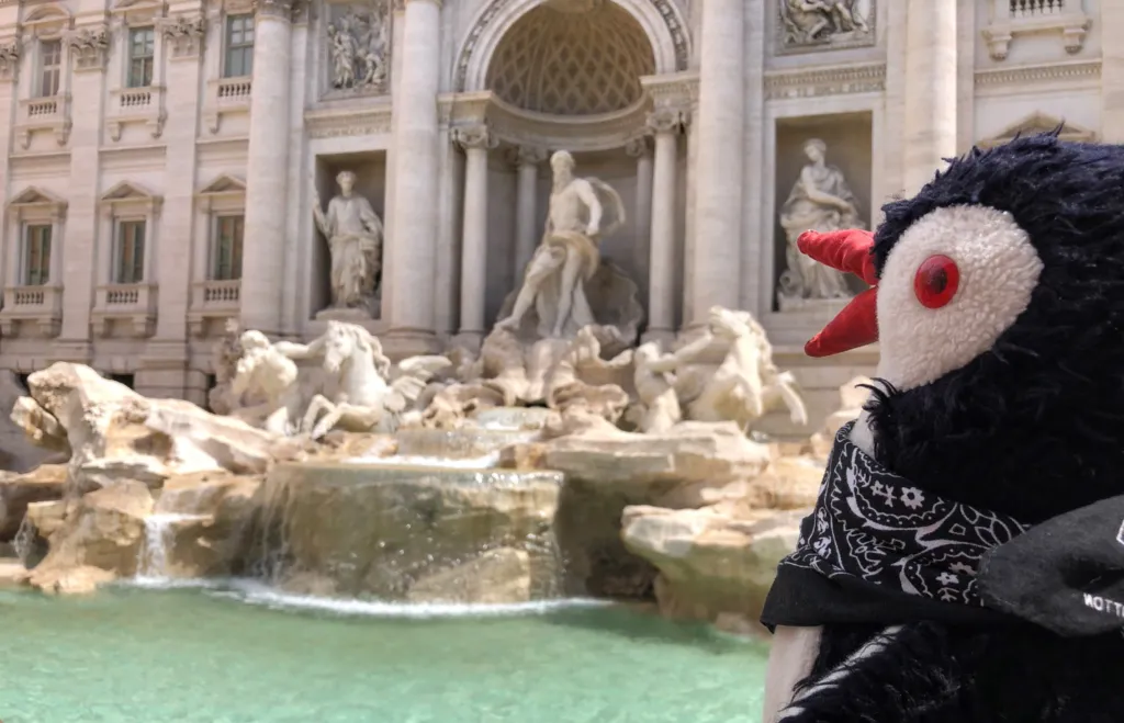 Plush penguin Miško wearing a bandana in front of the Trevi Fountain in Rome