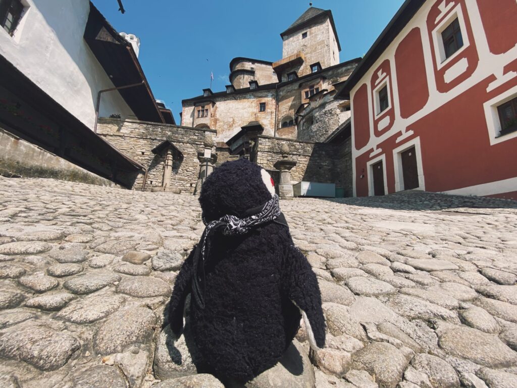 A plush penguin wearing a bandana sits on the cobblestone courtyard of Orava Castle