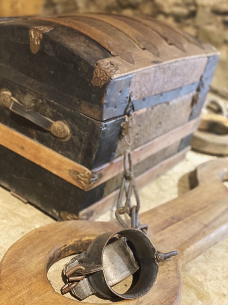 An antique chest sits near a wooden pillory with metal shackles at Stará Ľubovňa Castle