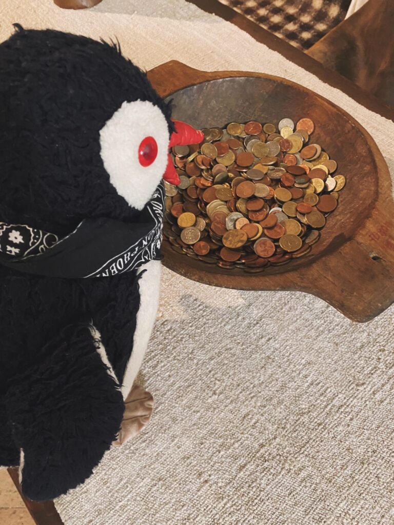 A plush penguin wearing a bandana looks at a wooden bowl of coins