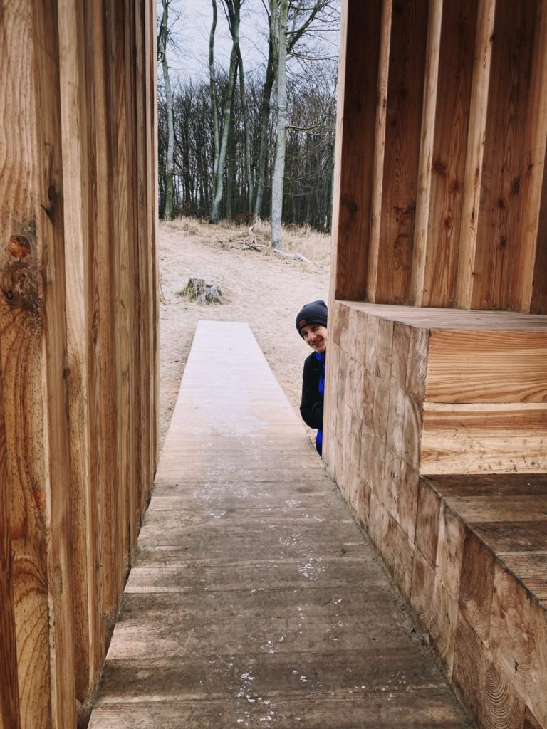 Person in a beanie peeking from behind a wooden structure in the forest
