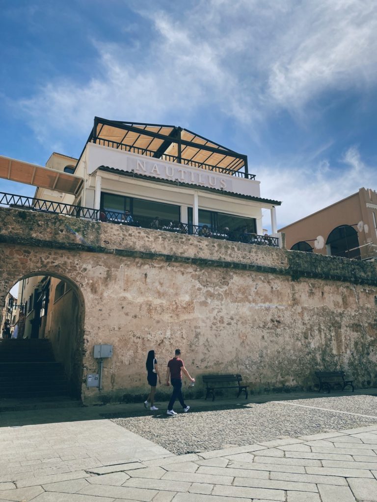 People walk past a stone wall beneath the Nautilus restaurant in Alghero