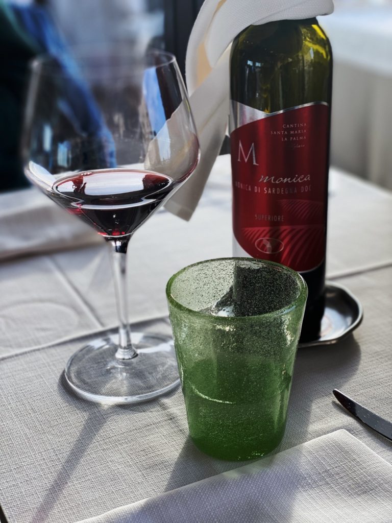 A bottle of red wine, a filled glass, and a green tumbler on an Alghero restaurant table