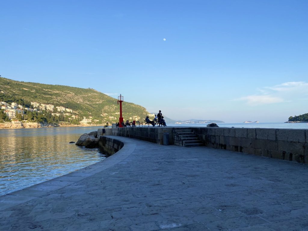 People sit on a stone pier with a red lighthouse in Dubrovnik