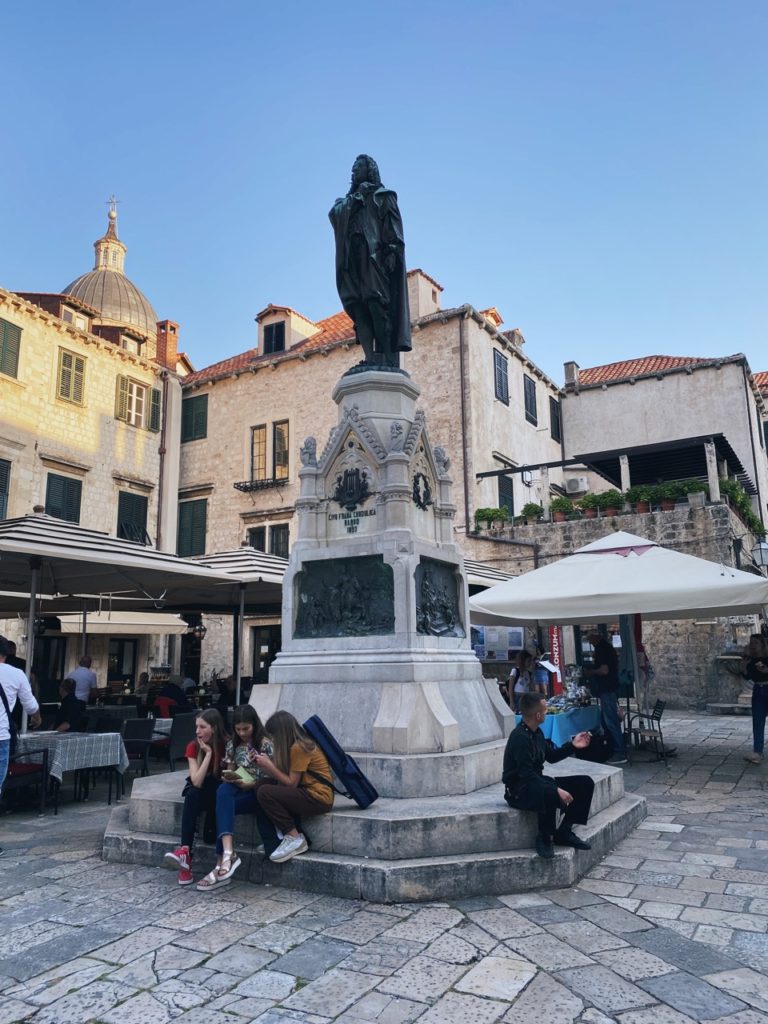 People sit on the base of the Gundulić Square monument in Dubrovnik