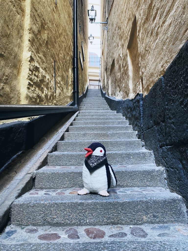 A plush penguin sits on stone steps in a narrow Stockholm alleyway