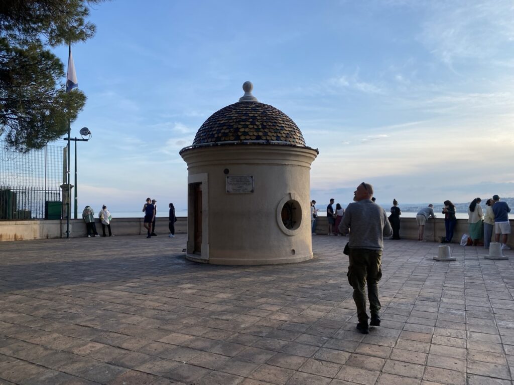 A small round building with a tiled dome stands on a paved terrace at Castle Hill in Nice as visitors look out