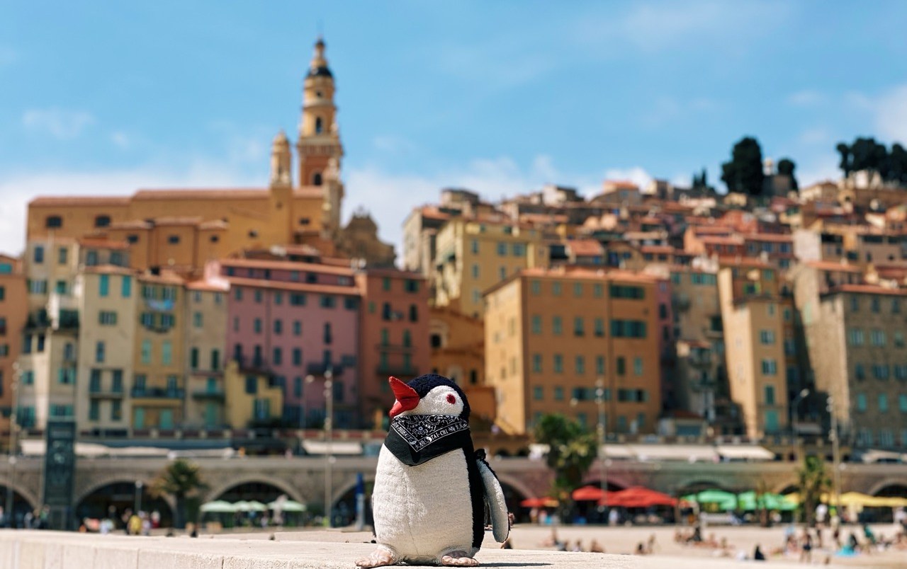 Miško the toy penguin on the beach in front of Menton's colourful old town, France