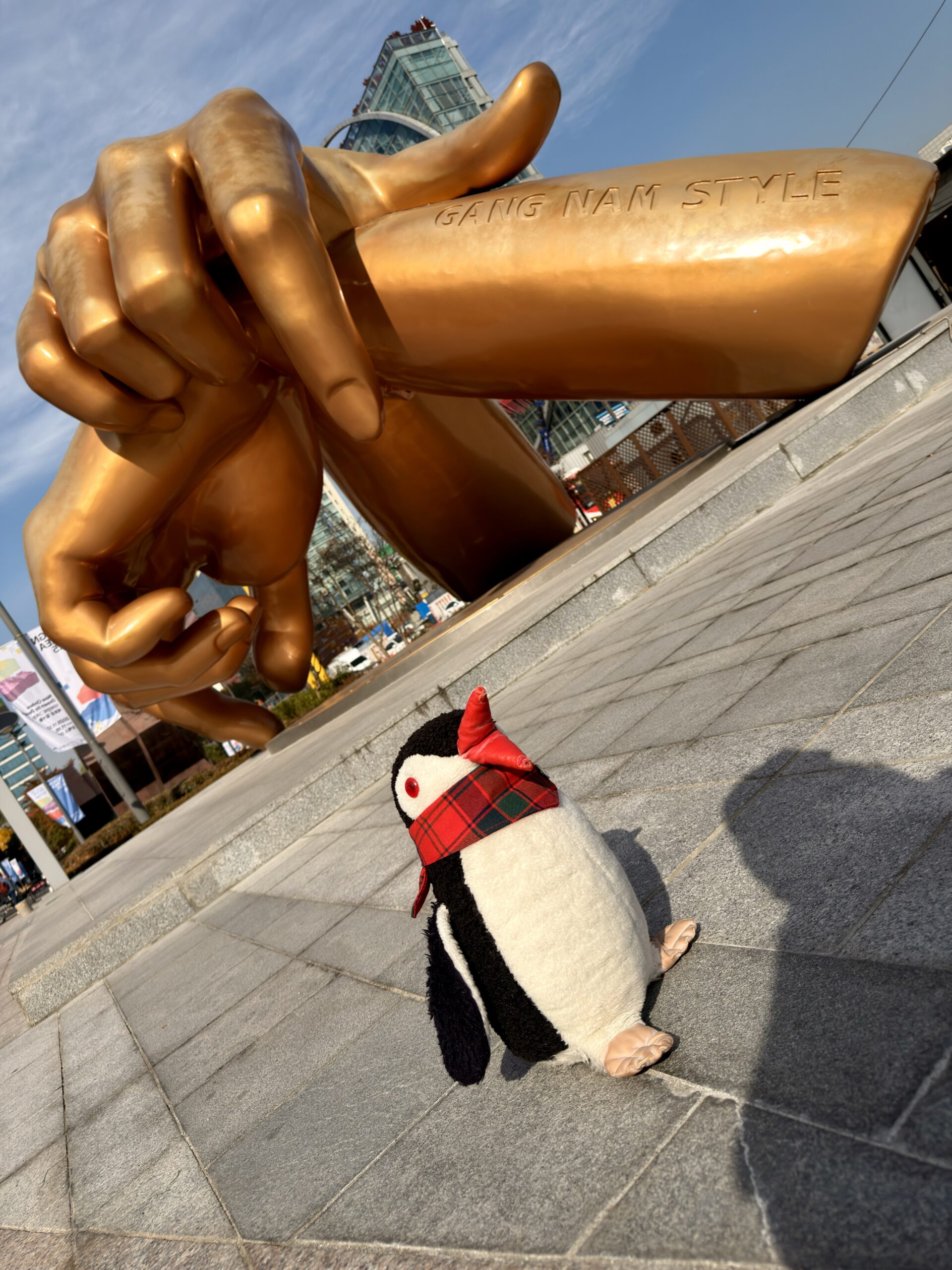 A plush penguin stands before the golden Gangnam Style hand sculpture in Seoul