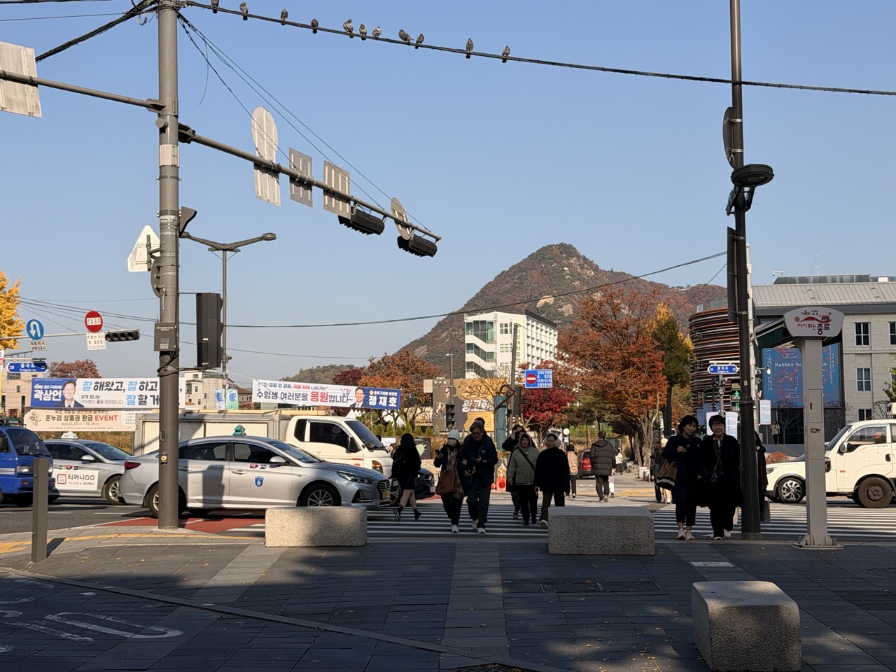 People walk across a Seoul street intersection with a mountain in the background