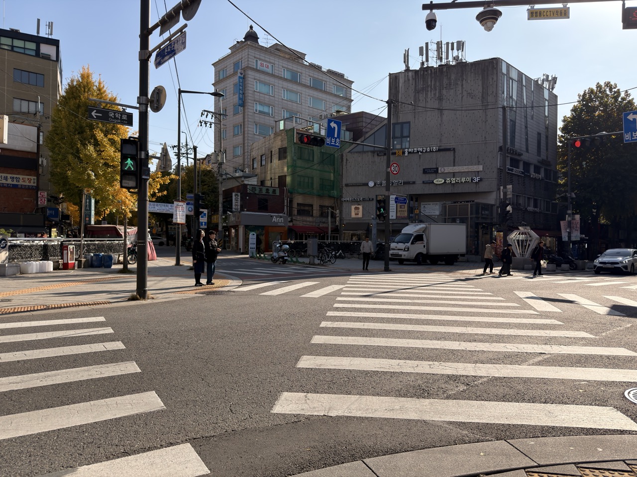Pedestrians wait at a sunny Seoul intersection with crosswalks and city buildings
