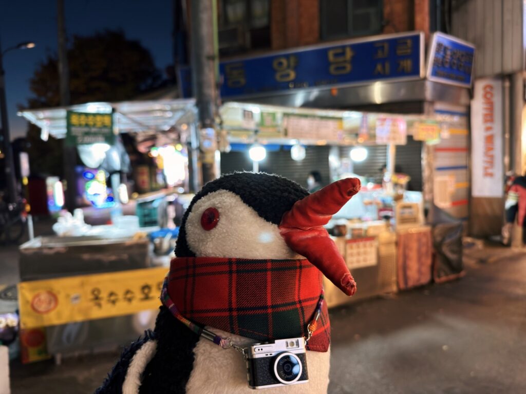 A plush penguin wearing a plaid scarf and toy camera at Gwangjang Market