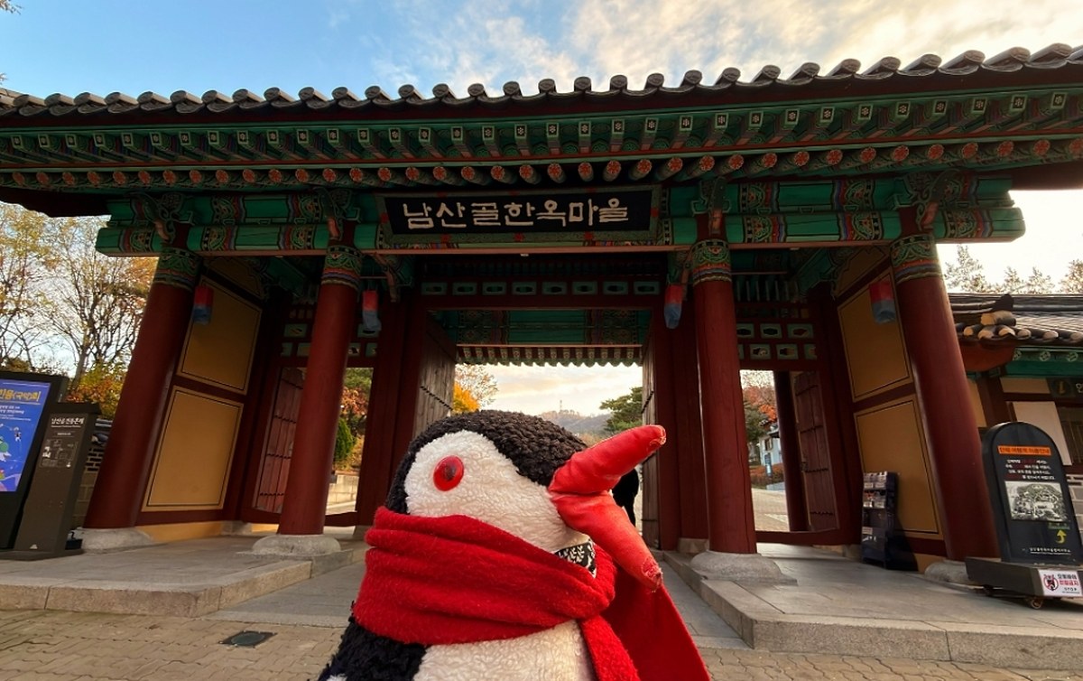 Miško the toy penguin with a red scarf stands before the traditional wooden gate at Namsangol Hanok Village