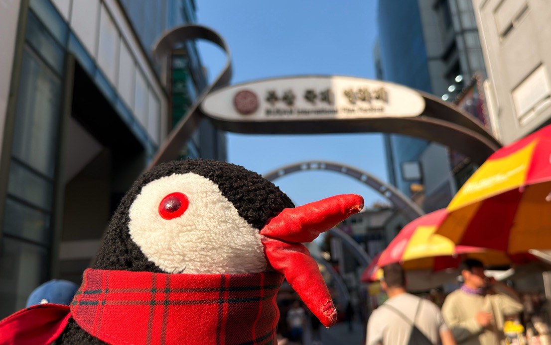 Miško the toy penguin in a red scarf under the BIFF Square sign in Busan, South Korea
