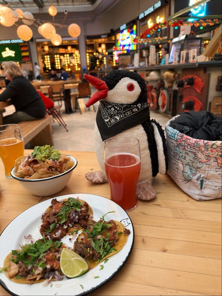 Stuffed penguin wearing a bandana sits at a table with tacos and drinks