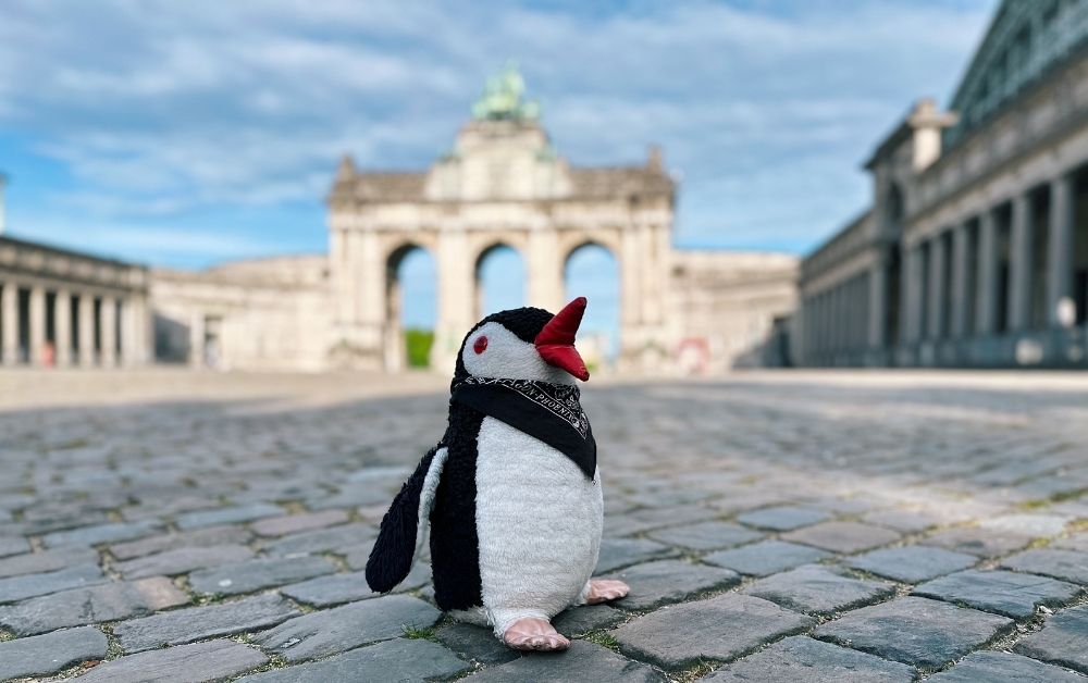 A plush penguin stands on the cobblestones before the Brussels Cinquantenaire arch