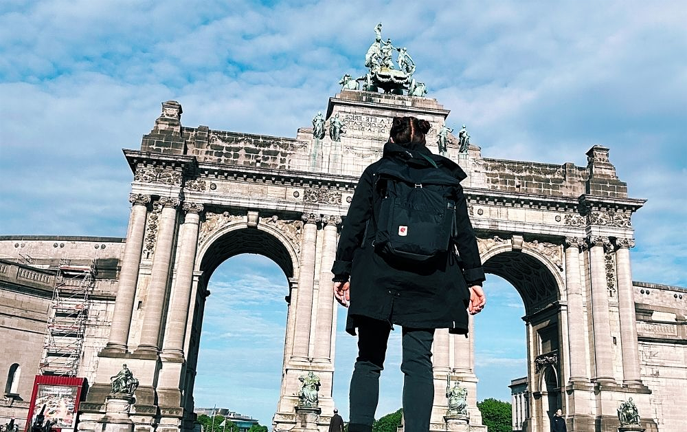 A woman with a black backpack looks up at a grand stone arch in Brussels topped with statues against a blue sky