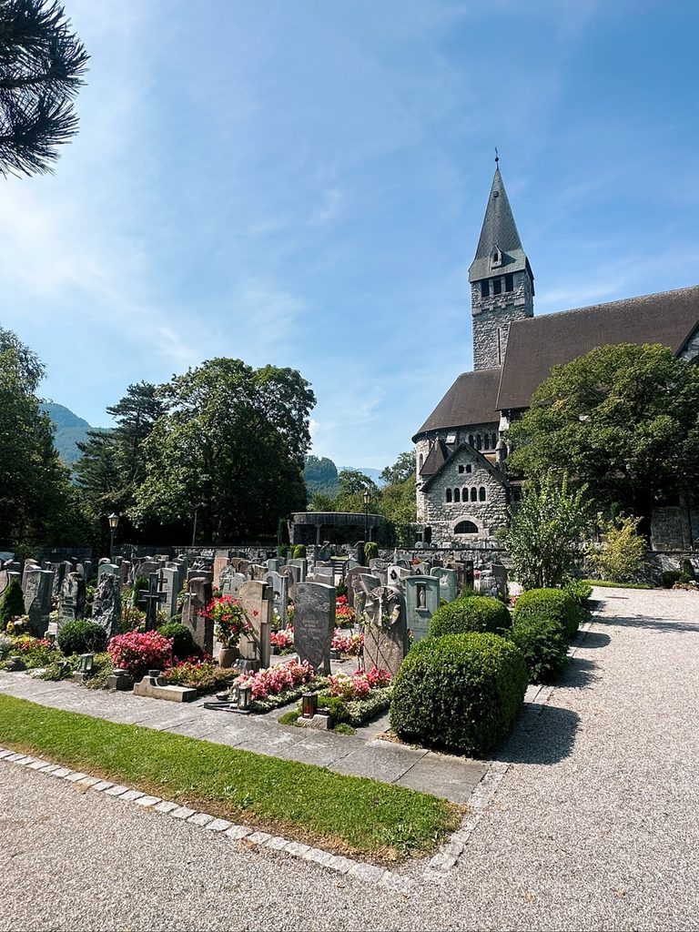 A cemetery with grey headstones and colorful flowers sits in front of a stone church with a tall steeple in Liechtenstein