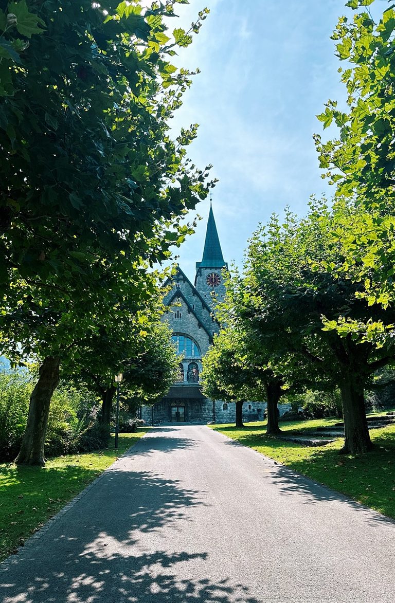A paved path leads through lush green trees toward a stone church with a tall green spire in Liechtenstein
