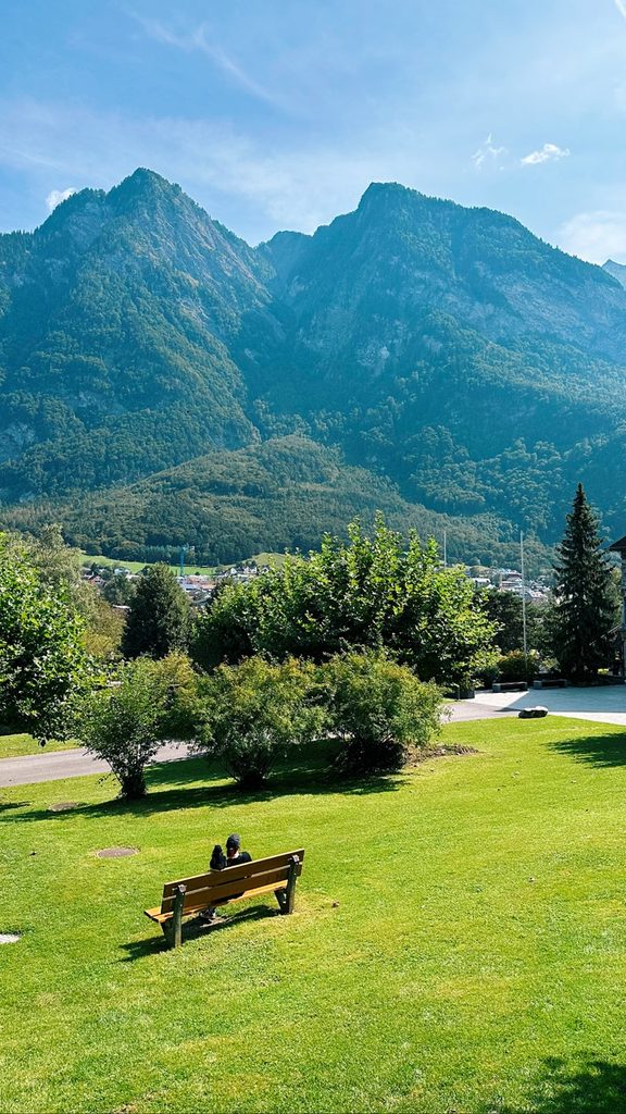 A person sits on a wooden bench in a grassy park facing green mountains and a town in Liechtenstein