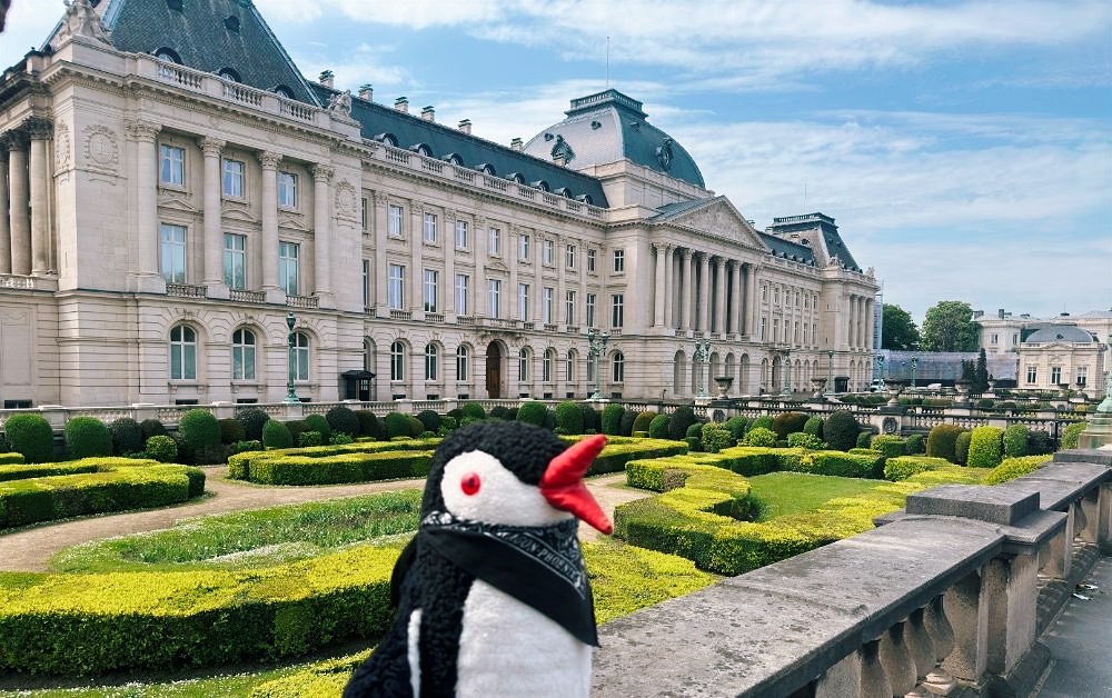 Miško the penguin with a red beak stands in front of a grand palace and gardens in Brussels