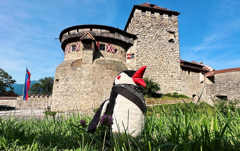 Miško the penguin sits in the grass in front of a stone castle in Liechtenstein