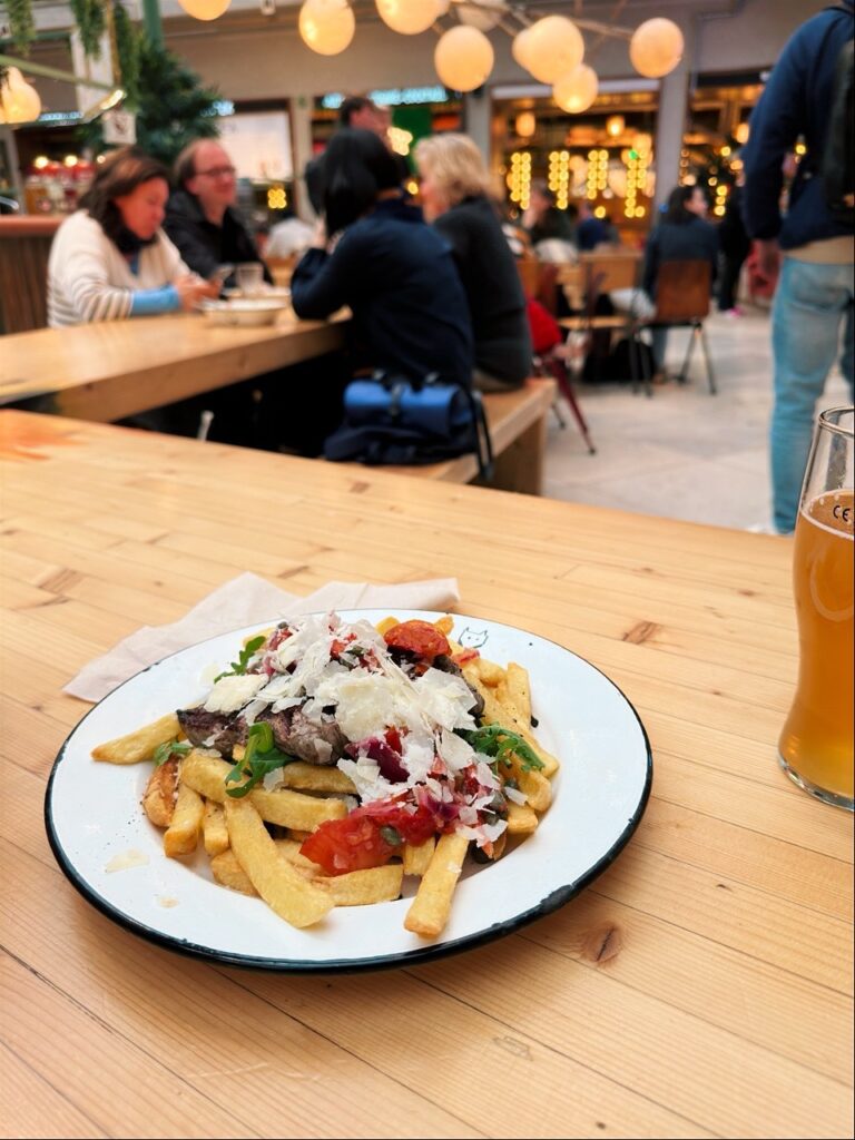 Plate of frites topped with steak, shaved cheese and salsa on a wooden table next to a beer glass in a busy Brussels restaurant
