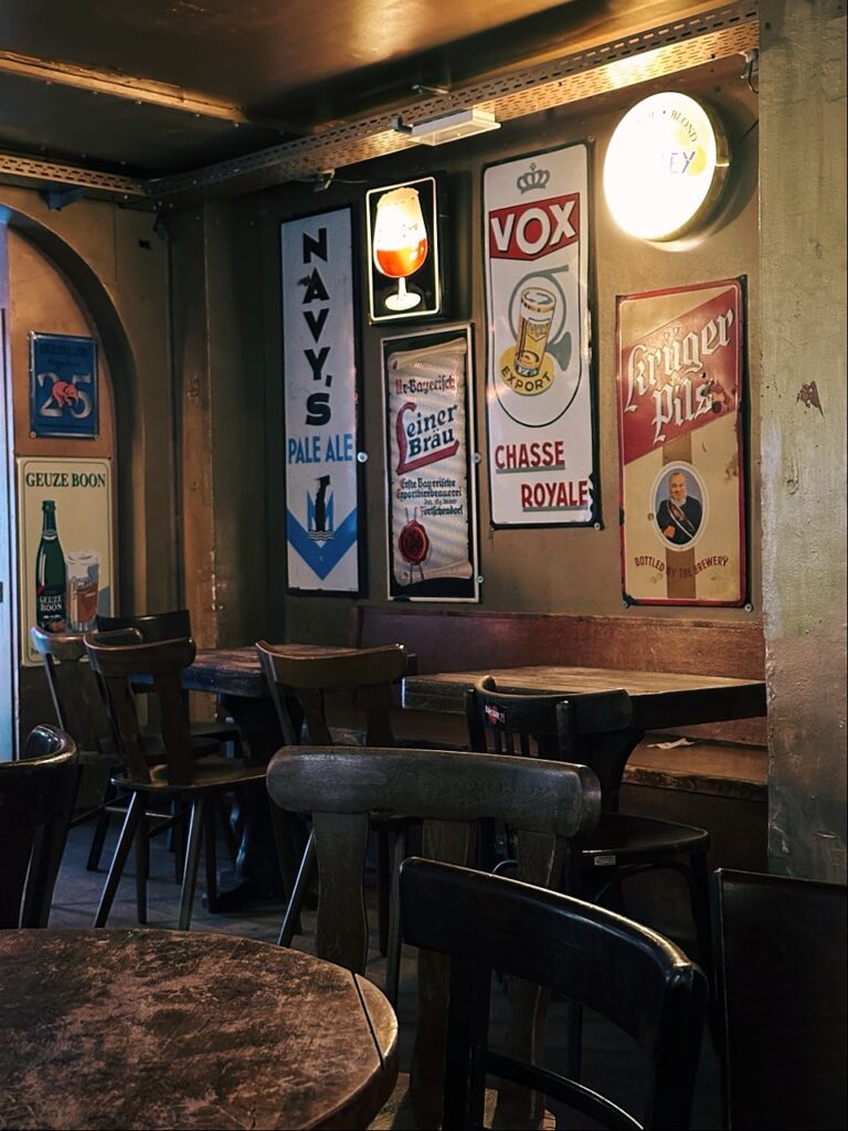 Wooden tables and chairs in a Brussels restaurant with vintage beer signs on the wall for Navy S Pale Ale and Vox Chasse Royale