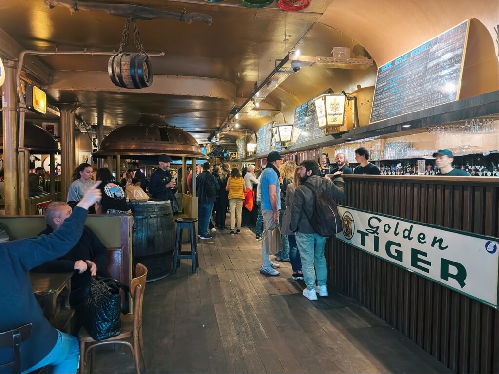 Crowded bar interior with patrons standing at a wooden counter displaying a Golden TIGER sign