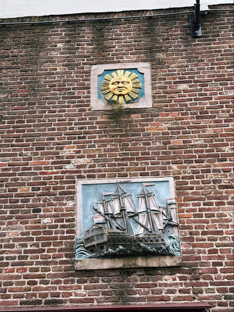 A brick wall in Amsterdam features a golden sun plaque above a relief of a sailing ship on waves