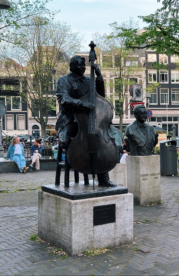 Bronze statue of a man playing a double bass stands next to a bust of Johnny Jordaan on a stone pedestal in Amsterdam