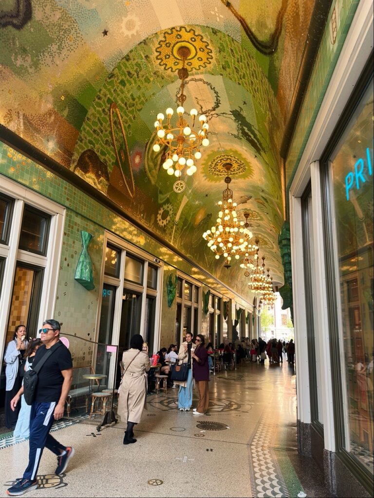 People walking through a covered arcade in Amsterdam featuring a green mosaic ceiling with gears and hanging chandeliers