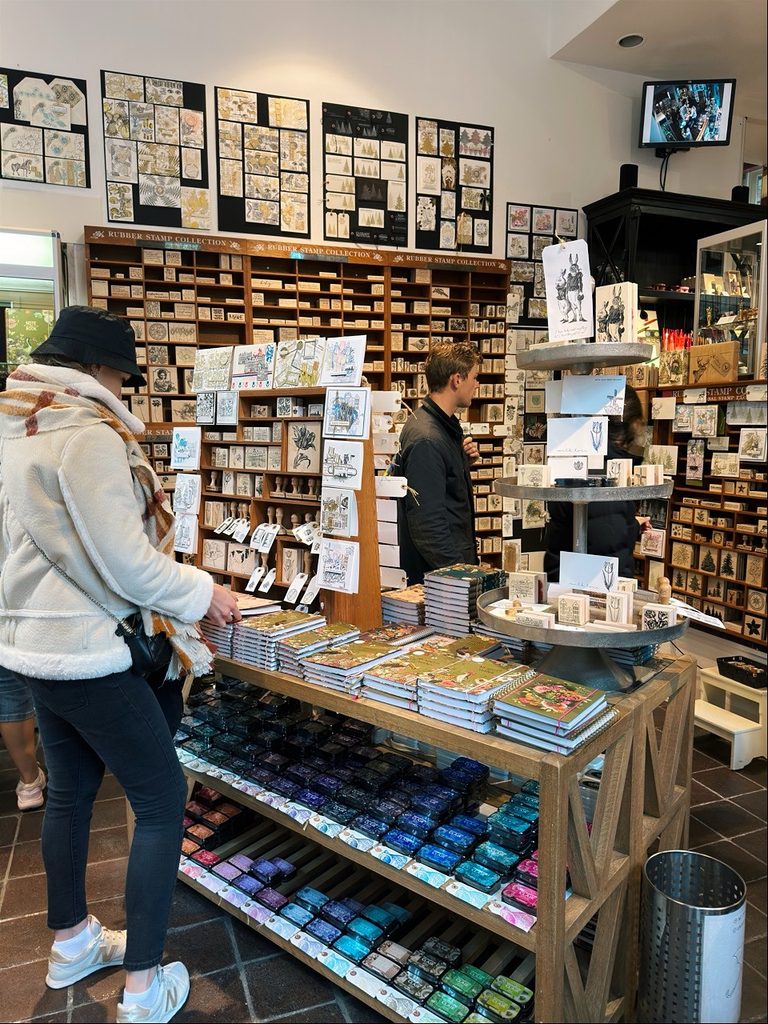A woman in a white jacket looks at notebooks and ink pads while a man stands near shelves labeled Rubber Stamp Collection