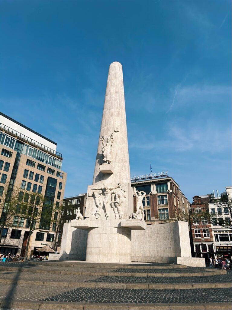 A tall white stone obelisk with sculptures at the base stands in a square in Amsterdam under a clear blue sky