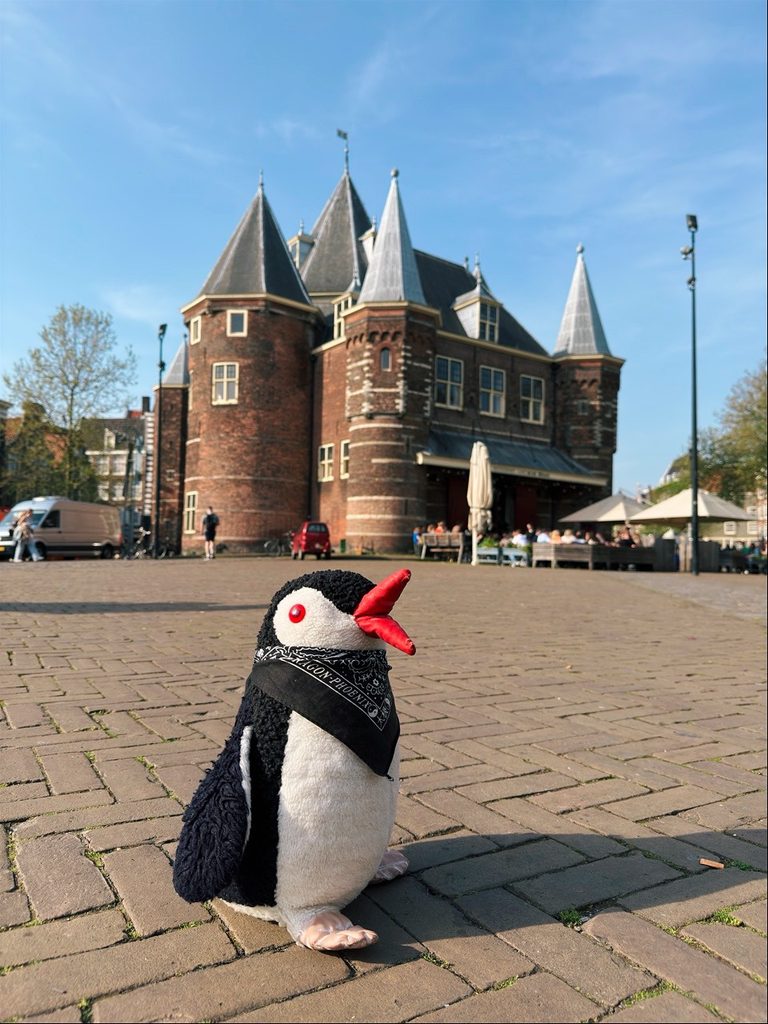 Miško the penguin wearing a black bandana sits on a brick pavement in front of a building with pointed towers in Amsterdam