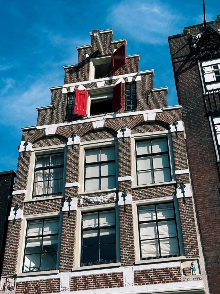 Tall brick building in Amsterdam with a stepped gable, red shutters, hoist hooks, and tiles reading 1649 and D' BROUWER