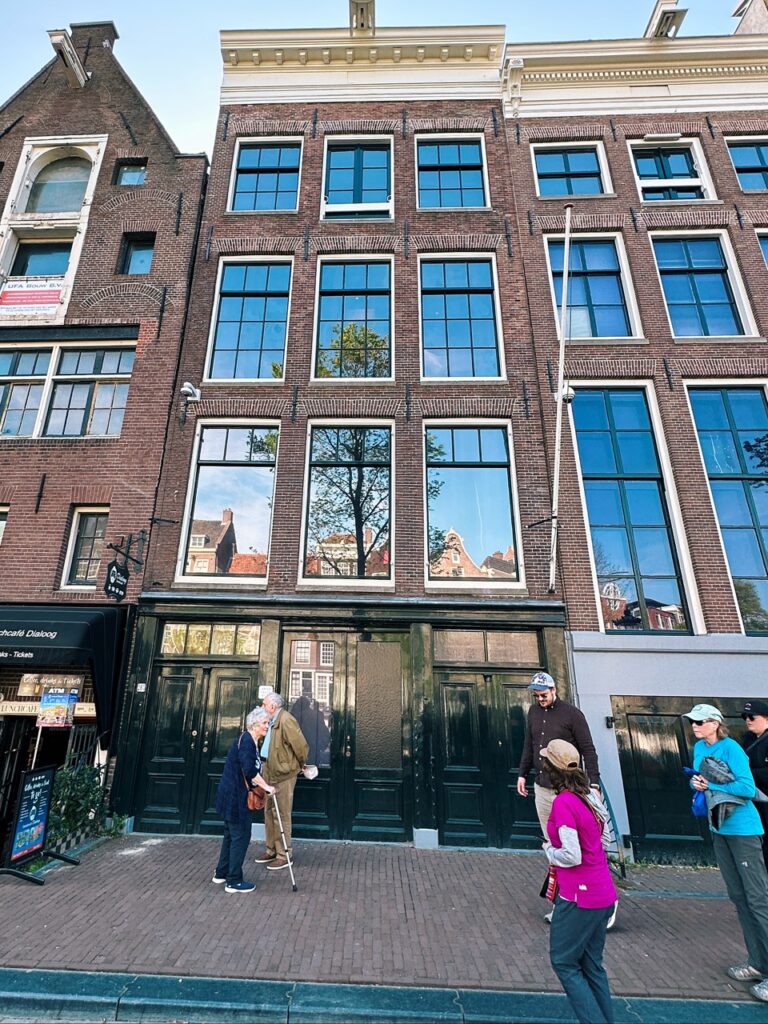 The brick facade of the Anne Frank House features tall windows and dark green doors with several visitors standing on the sidewalk