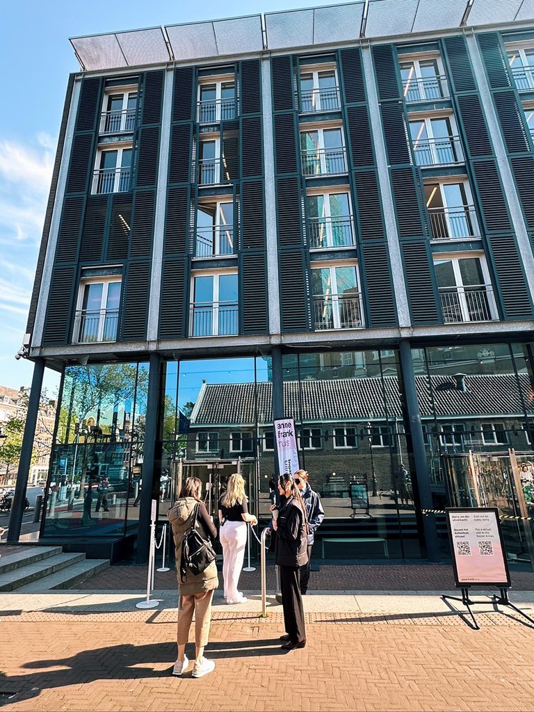 Visitors stand outside the glass entrance of the Anne Frank House, a modern building with dark cladding and rows of windows