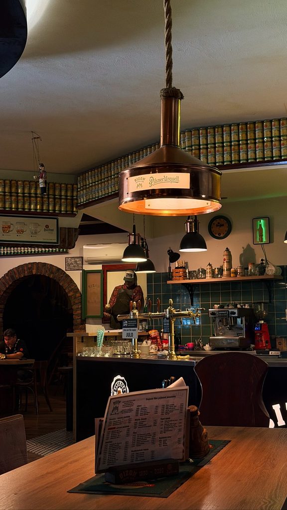 Bizon Pub Bratislava interior with a bartender, walls lined with beer cans, a Pilsner Urquell lamp, and a menu on a table
