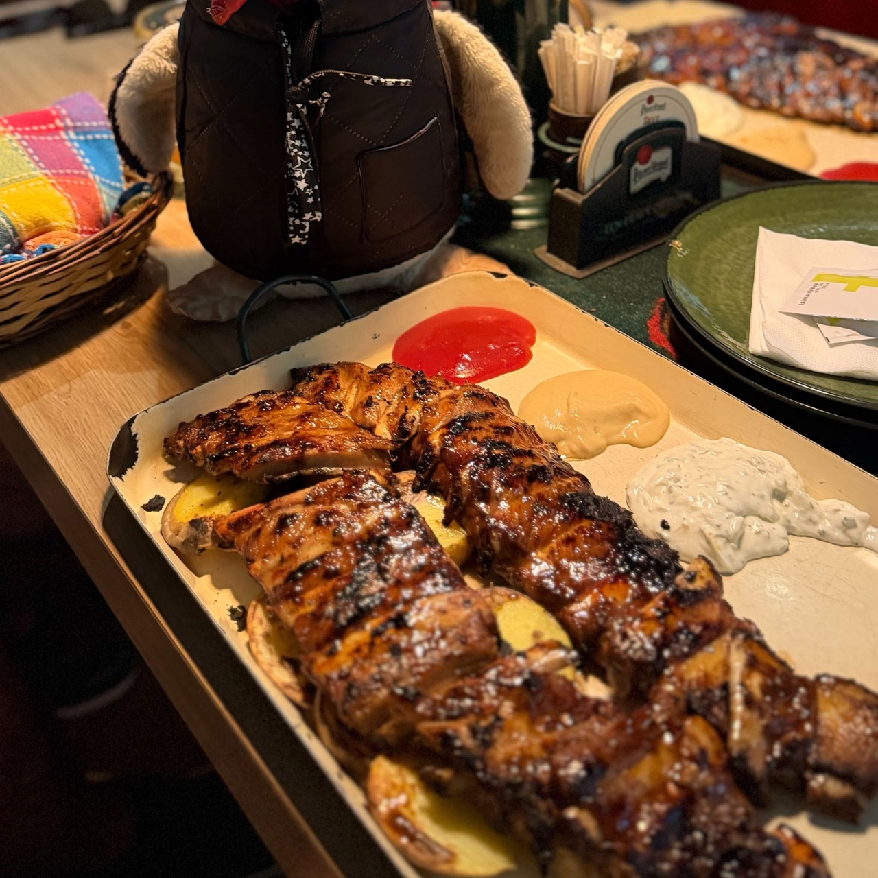 Large platter of grilled ribs with potato slices and three dipping sauces while Miško the knitted penguin stands in the background