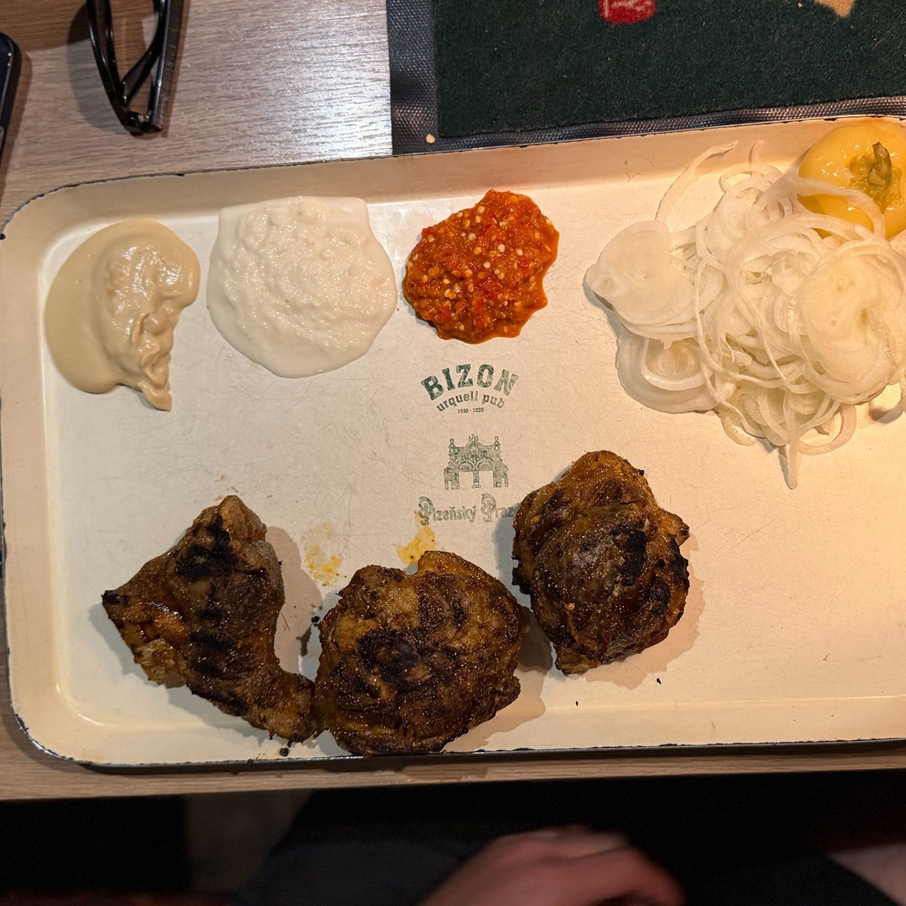 Three pieces of grilled meat on a white tray marked BIZON urquell pub, served with three sauces, sliced onions and a lemon wedge