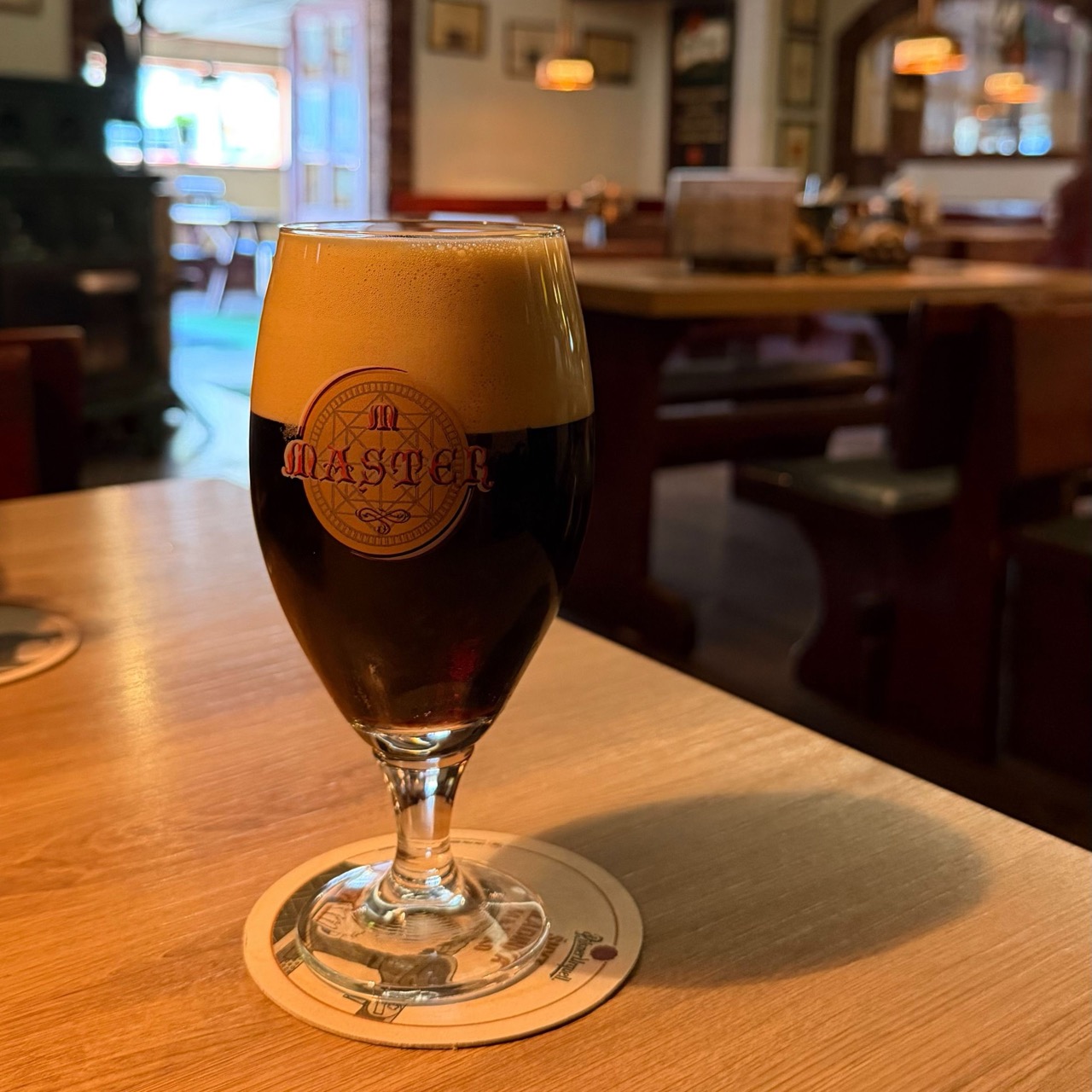 A glass of dark beer with the Master logo sits on a coaster at a wooden table in Bizon Pub Bratislava