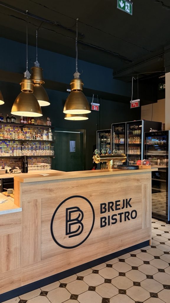 The wooden bar at Brejk Bistro Prešov features the BREJK BISTRO logo under gold pendant lights with shelves of drinks behind