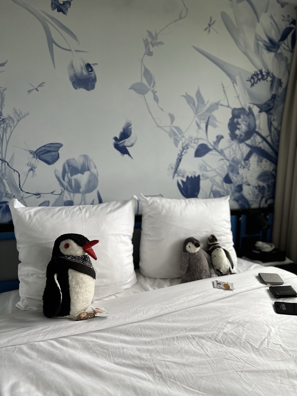 Miško and two smaller stuffed penguins sit on a white bed with blue floral wallpaper at Dutch Design Hotel Vondelpark Amsterdam