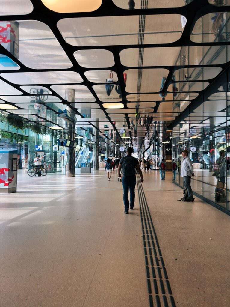 A man with a backpack walks through a modern Amsterdam station hall featuring a unique black grid ceiling and glass walls