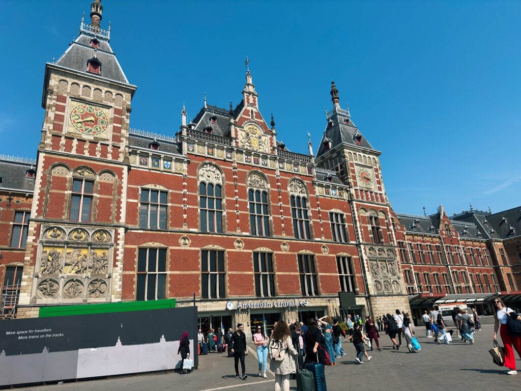 Travellers walk in front of the red brick Amsterdam Centraal station building under a blue sky