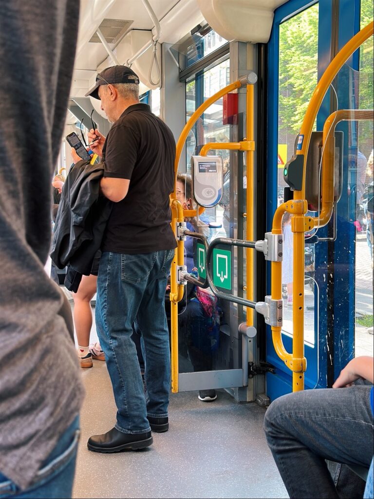 A man in a black cap stands inside an Amsterdam tram looking at his phone near yellow handrails and a card reader