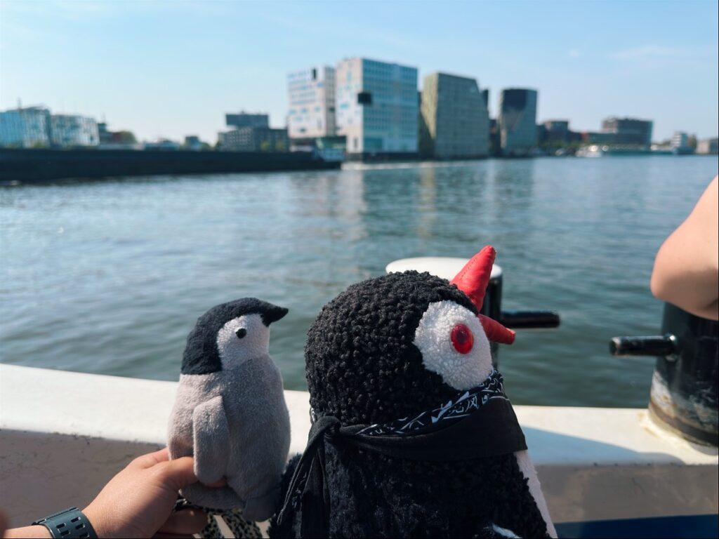 Miško and a smaller plush penguin held up on a boat with Amsterdam buildings visible across the water